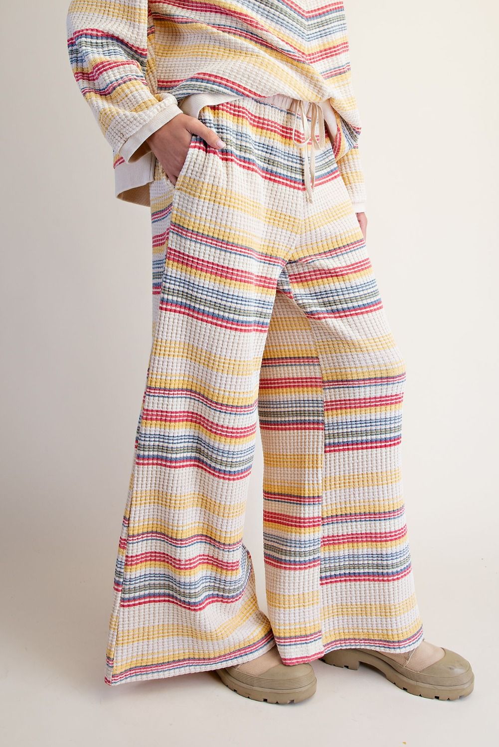 A person wearing wide-leg striped pants and a matching striped top, standing against a plain light beige studio background. The clothing has horizontal multicolor stripes including yellow, red, blue, gray, and white, and the person is wearing beige shoes.