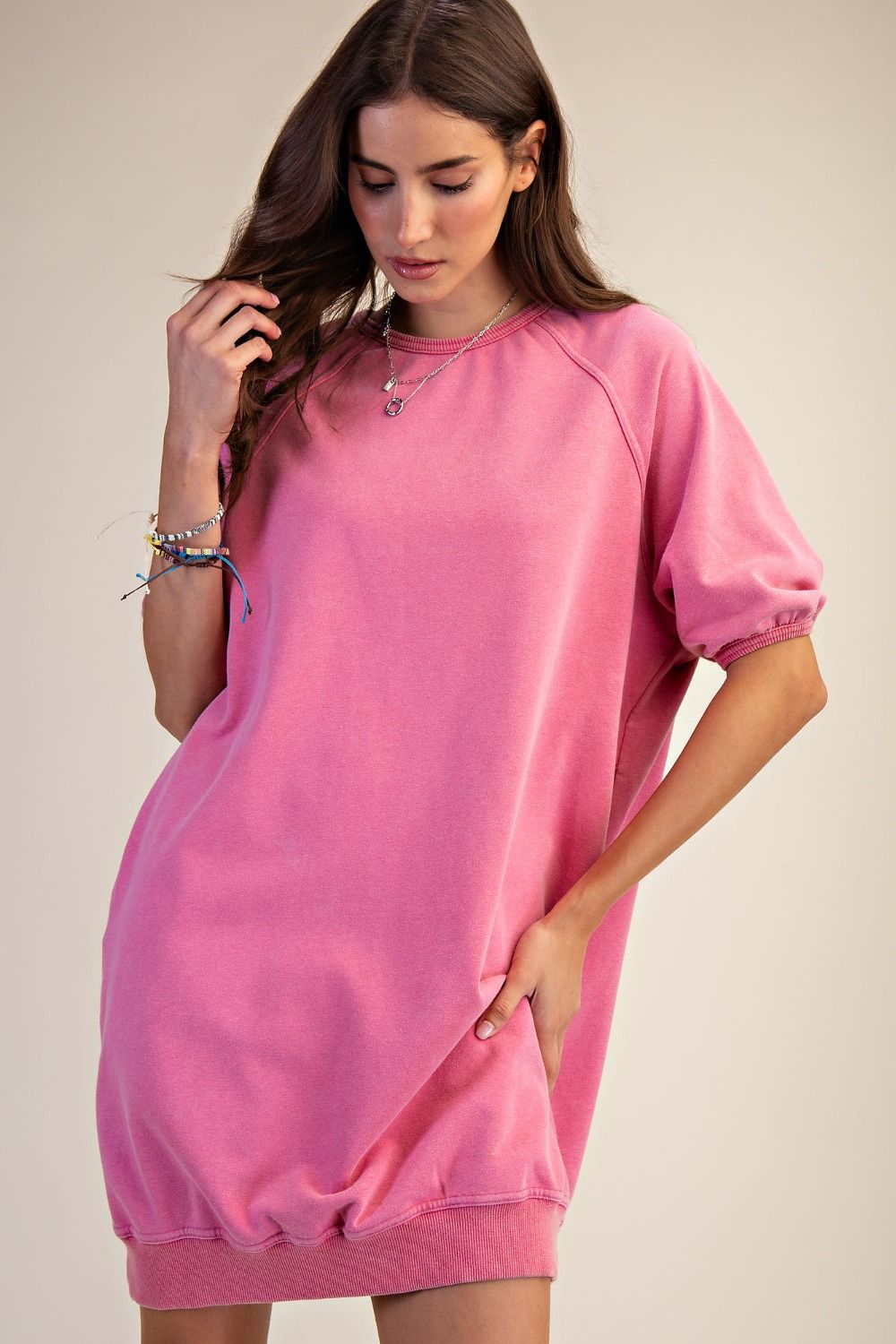 A woman wearing an oversized pink sweatshirt dress with short puff sleeves and a relaxed fit, posed against a plain beige studio background.