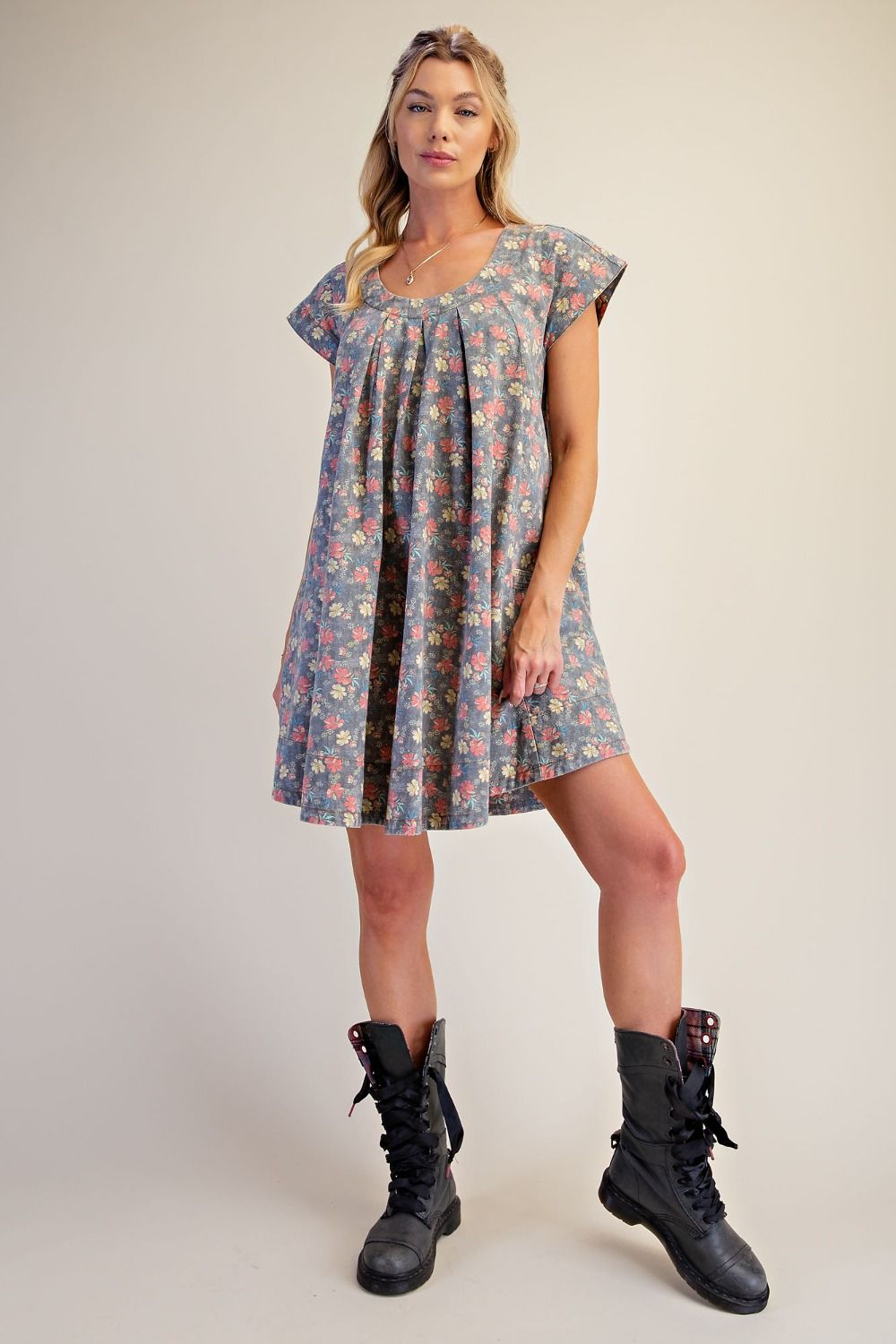 A woman modeling a short flutter-sleeve dress with a blue-gray base and a dense multicolor floral print, paired with black lace-up combat boots, against a solid light beige studio background.