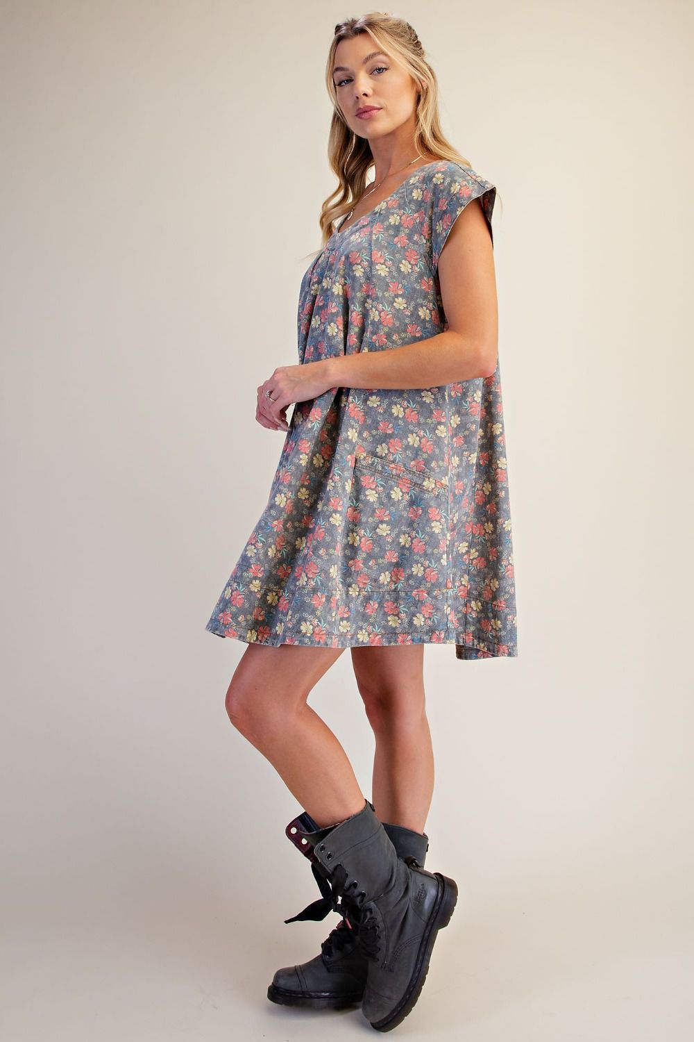 A woman modeling a short, sleeveless V-neck dress with a muted blue-gray base and a small multicolor floral print, paired with black lace-up ankle boots against a plain beige studio background.