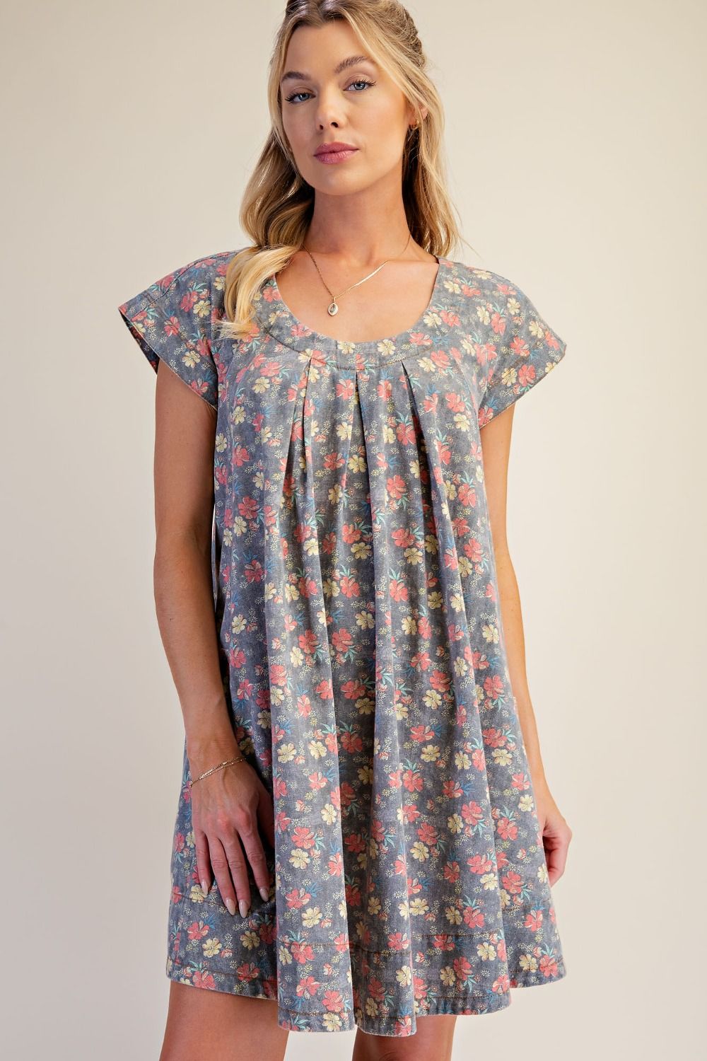 A woman wearing a short-sleeve floral print dress with a pleated bodice and a flared skirt, shown against a plain beige studio background.