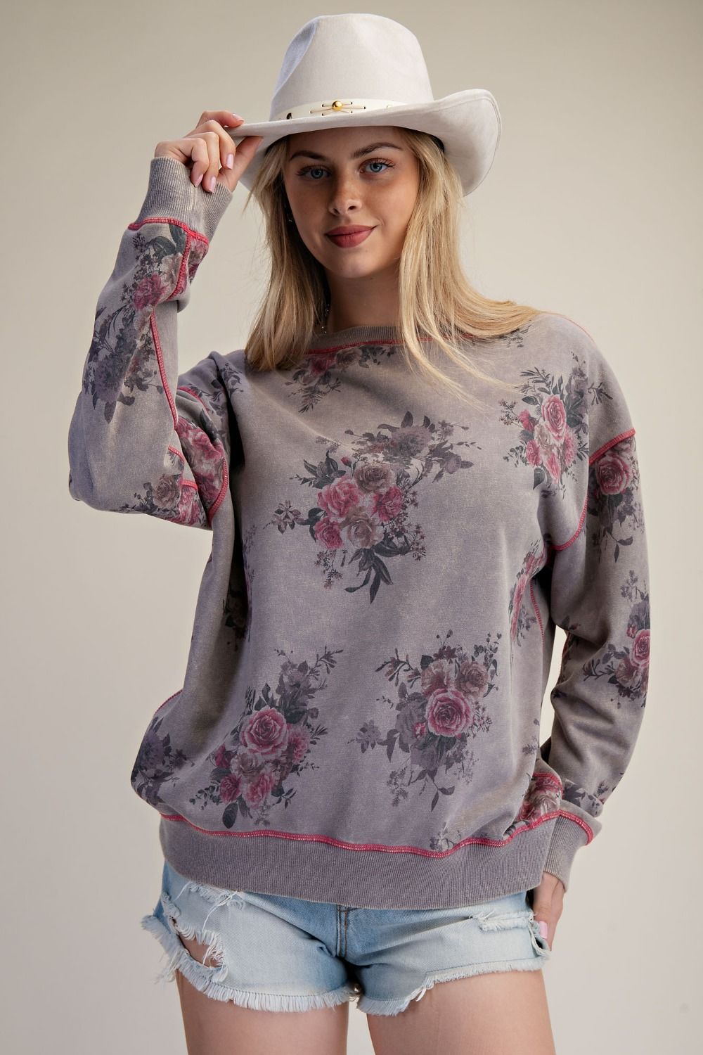 A woman wearing a gray crewneck sweatshirt with a pink and green floral pattern, styled with a white cowboy hat and denim shorts against a neutral beige background.
