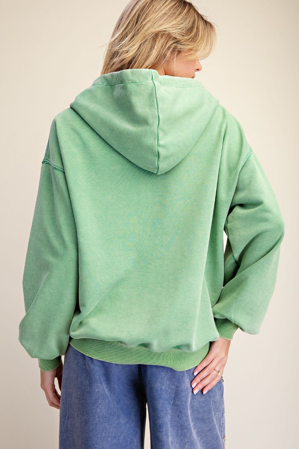 A woman shown from the back wearing an oversized sage green hooded sweatshirt and blue jeans against a plain beige studio background.
