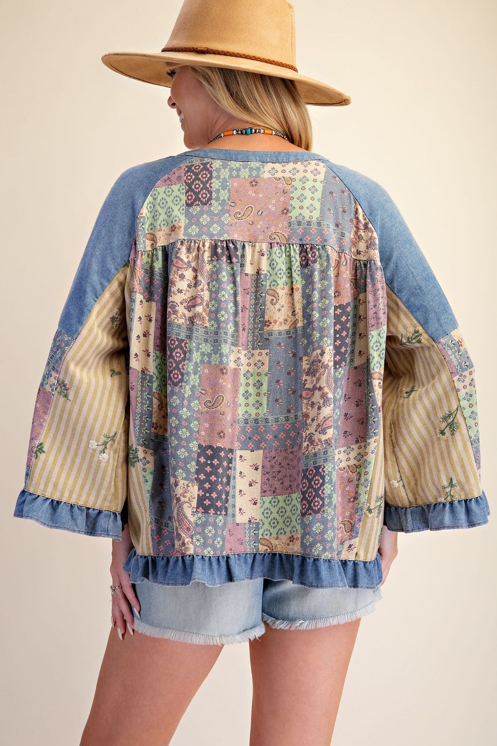 A woman shown from the back wearing a bohemian-style patchwork denim top with long bell sleeves and ruffled trim, paired with denim shorts and a straw hat against a solid beige background.