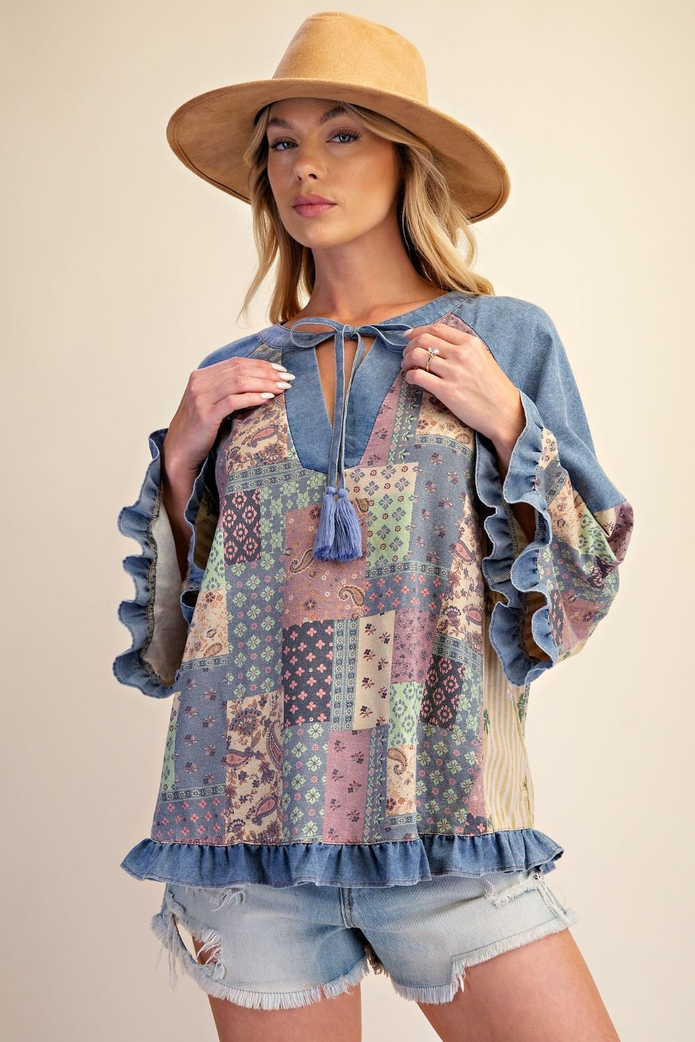 A woman wearing a bohemian-style patchwork top with blue denim ruffle trim, a tie neckline, and a straw hat, posing against a solid beige background.