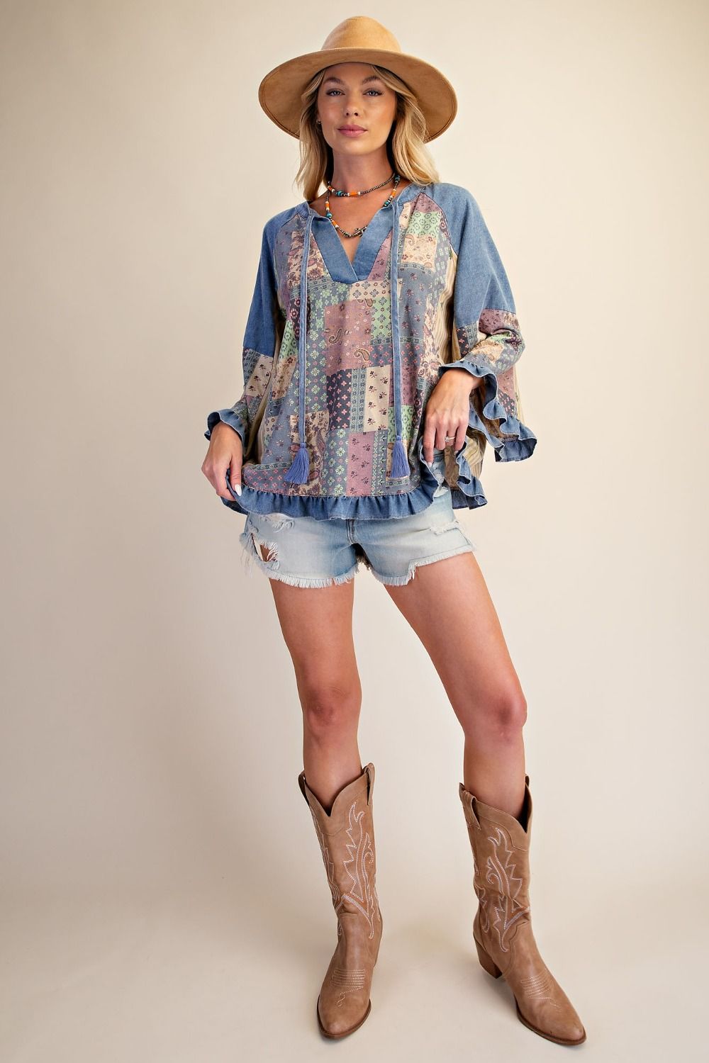 A woman modeling a bohemian-style blue patchwork blouse with ruffled sleeves, paired with denim shorts, a tan hat, and brown cowboy boots, against a solid beige studio background.