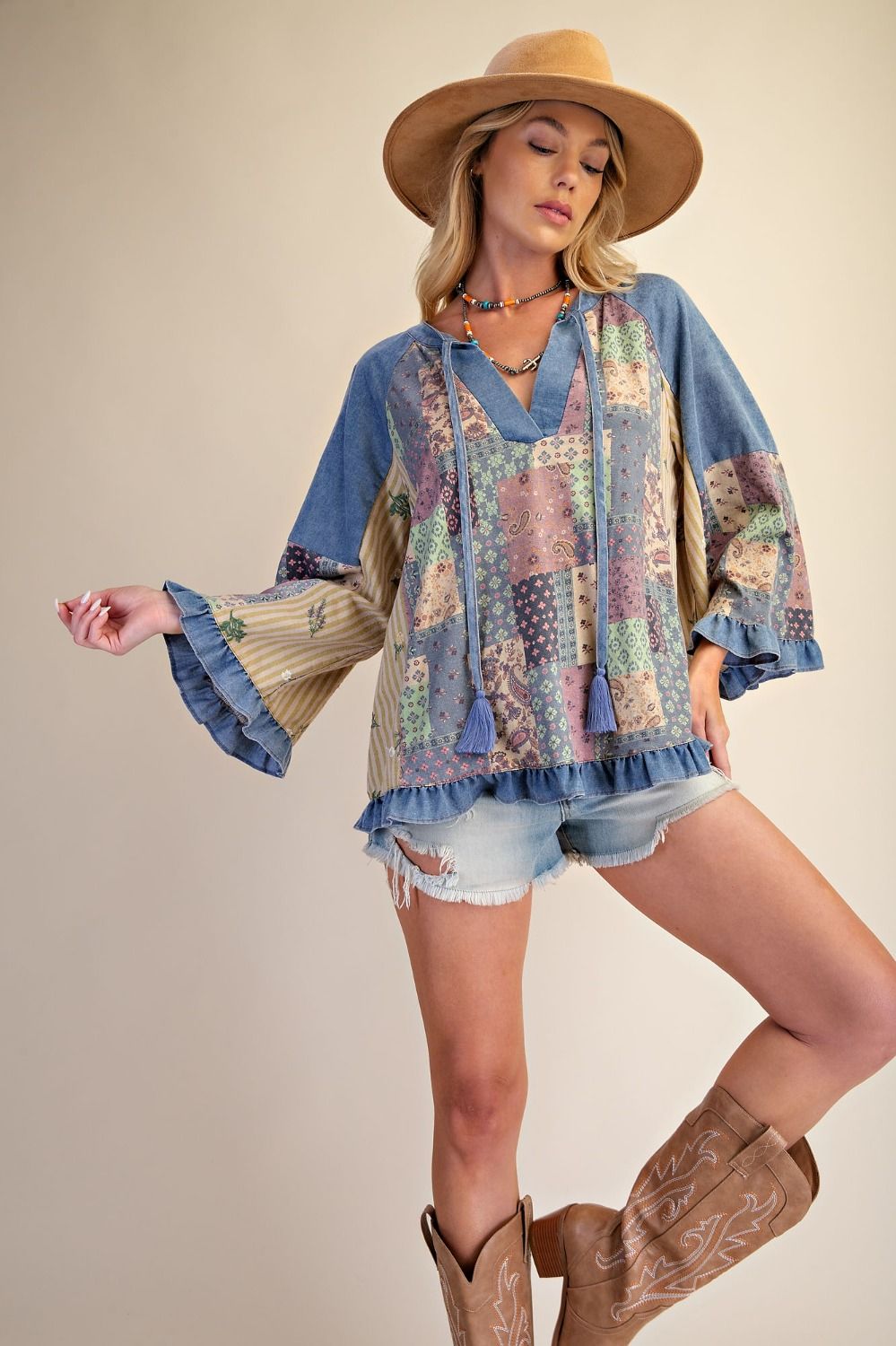 A woman modeling a bohemian-style blue patchwork tunic top with tassel ties, paired with denim shorts, a tan wide-brim hat, and brown cowboy boots against a plain beige studio background.