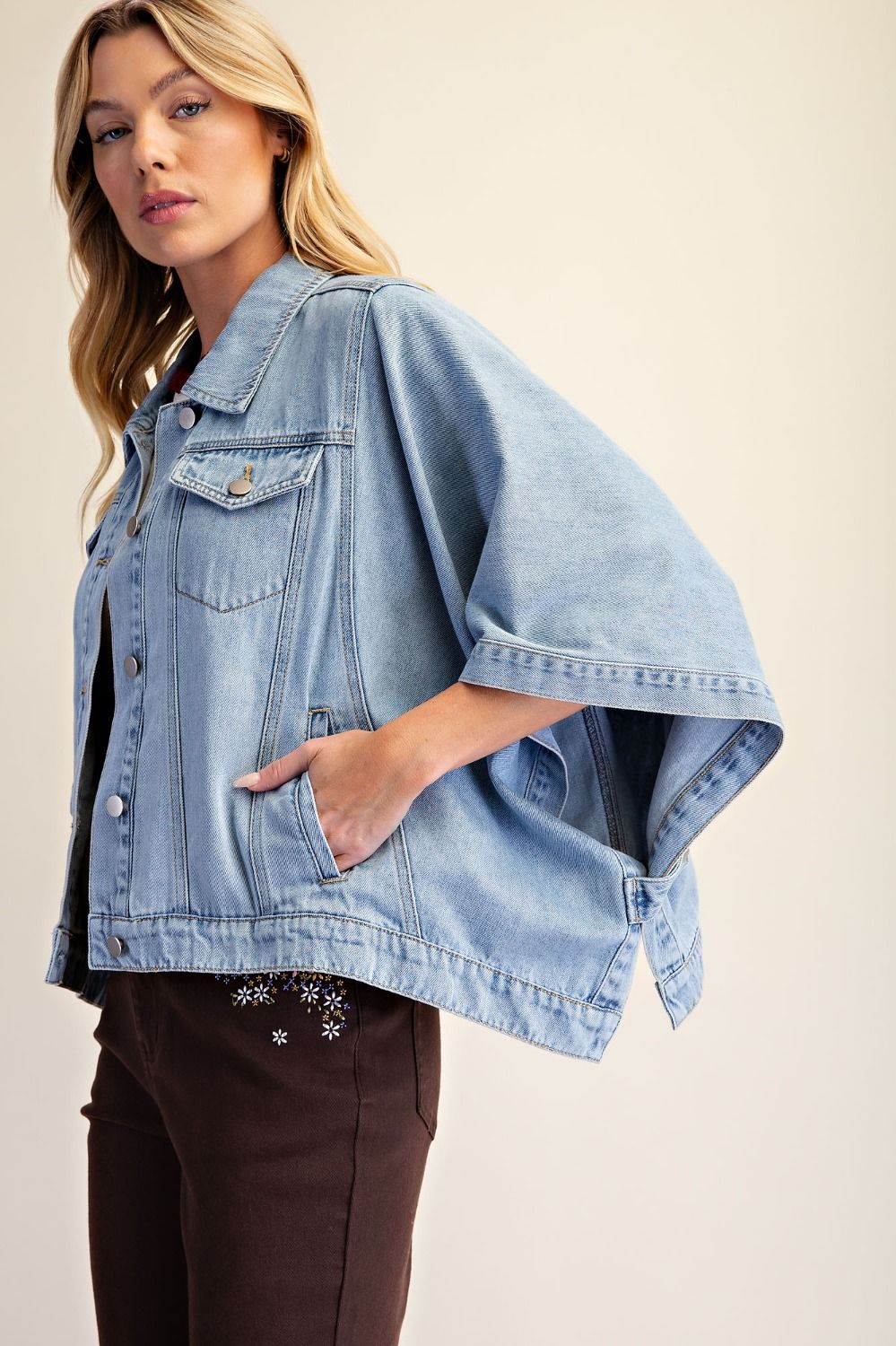 A woman wearing a light blue cropped denim jacket with wide short sleeves, button front, chest flap pockets, and side pockets, paired with dark brown pants against a plain beige studio background.