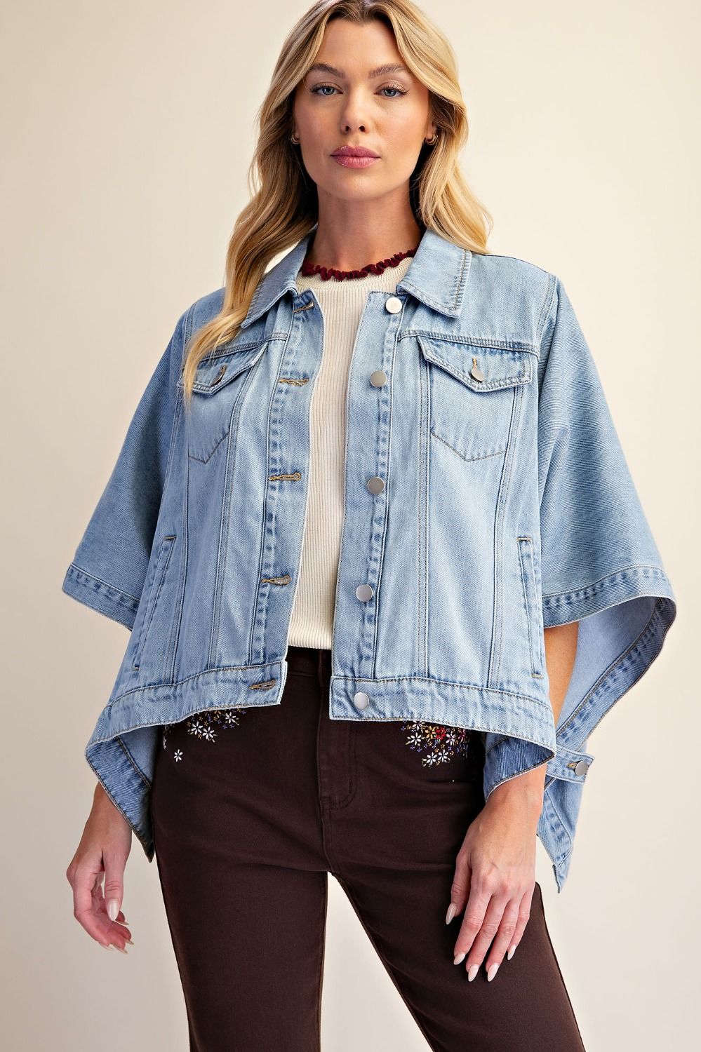 A woman modeling a light-wash denim jacket with wide short sleeves, worn over a cream top and dark brown pants, against a solid beige background.