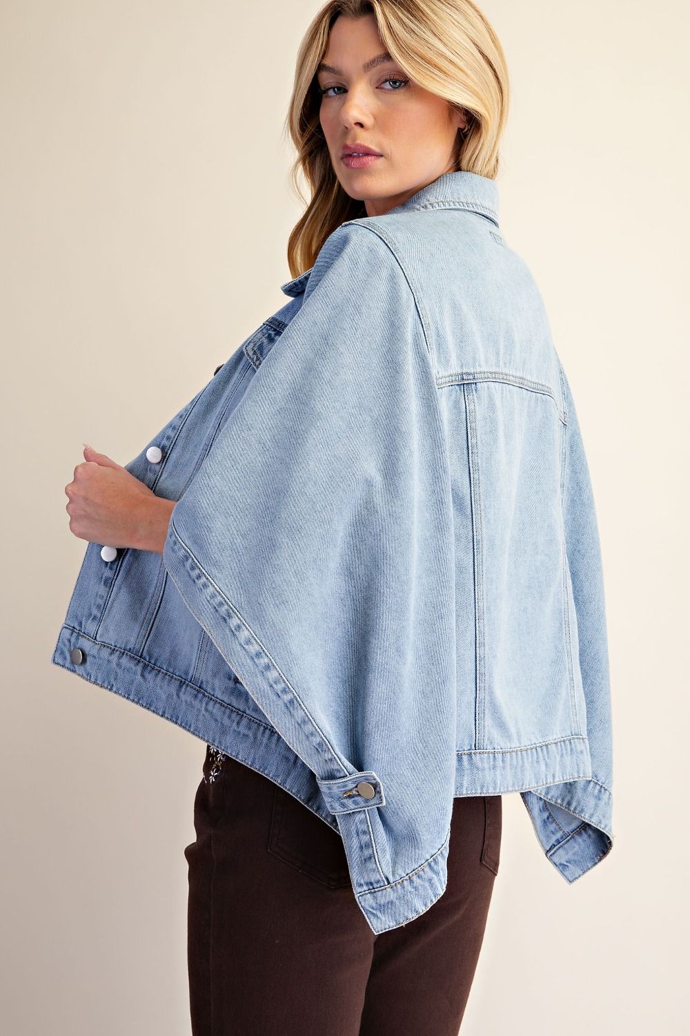 A woman wearing a light blue cropped denim poncho jacket with wide draped sleeves, shown from a three-quarter back angle against a plain beige studio background.