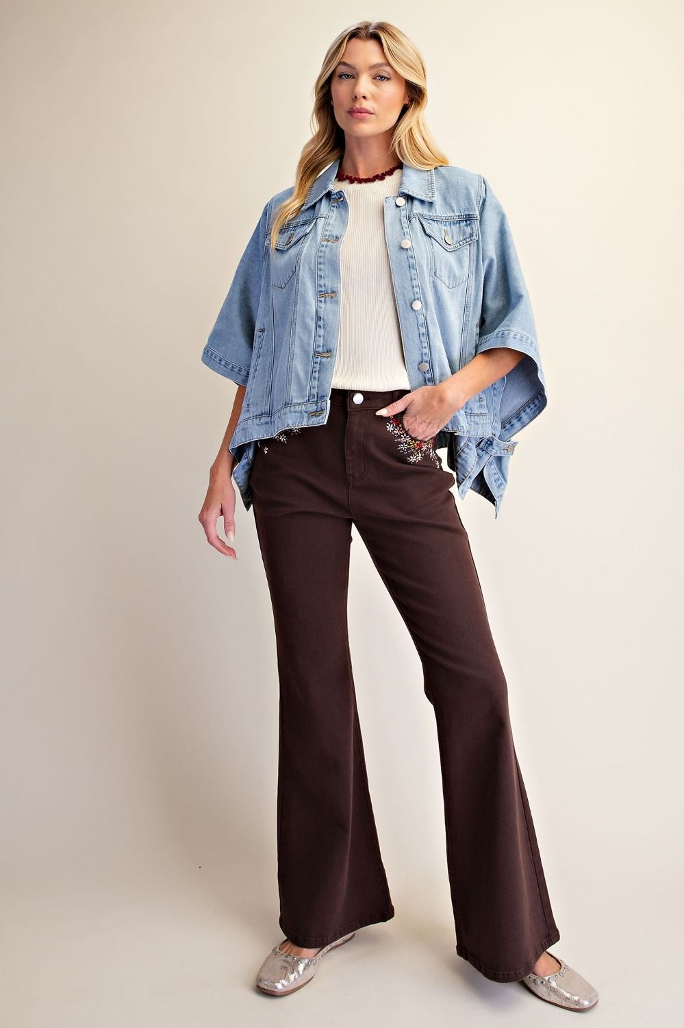 A woman modeling a light blue cropped denim jacket with wide sleeves, paired with dark brown flared pants, a white ribbed top, and silver flats, against a plain beige studio background.