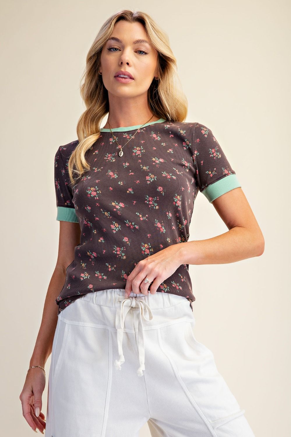 A woman modeling a short-sleeve ringer t-shirt with a dark brown floral print and mint green trim on the collar and cuffs, paired with white drawstring pants against a solid beige background.