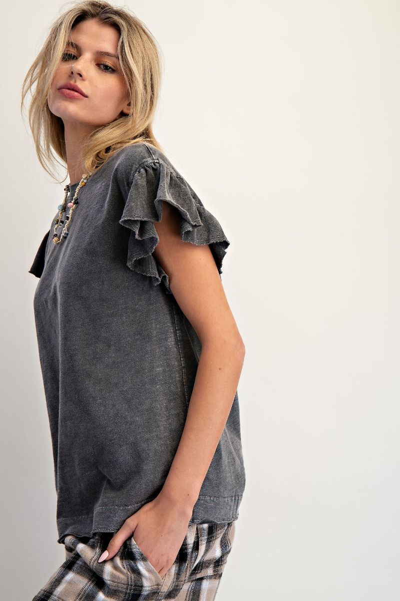 A woman wearing a dark gray washed short-sleeve top with ruffled detailing on the sleeves, paired with plaid pants, photographed against a plain off-white background.