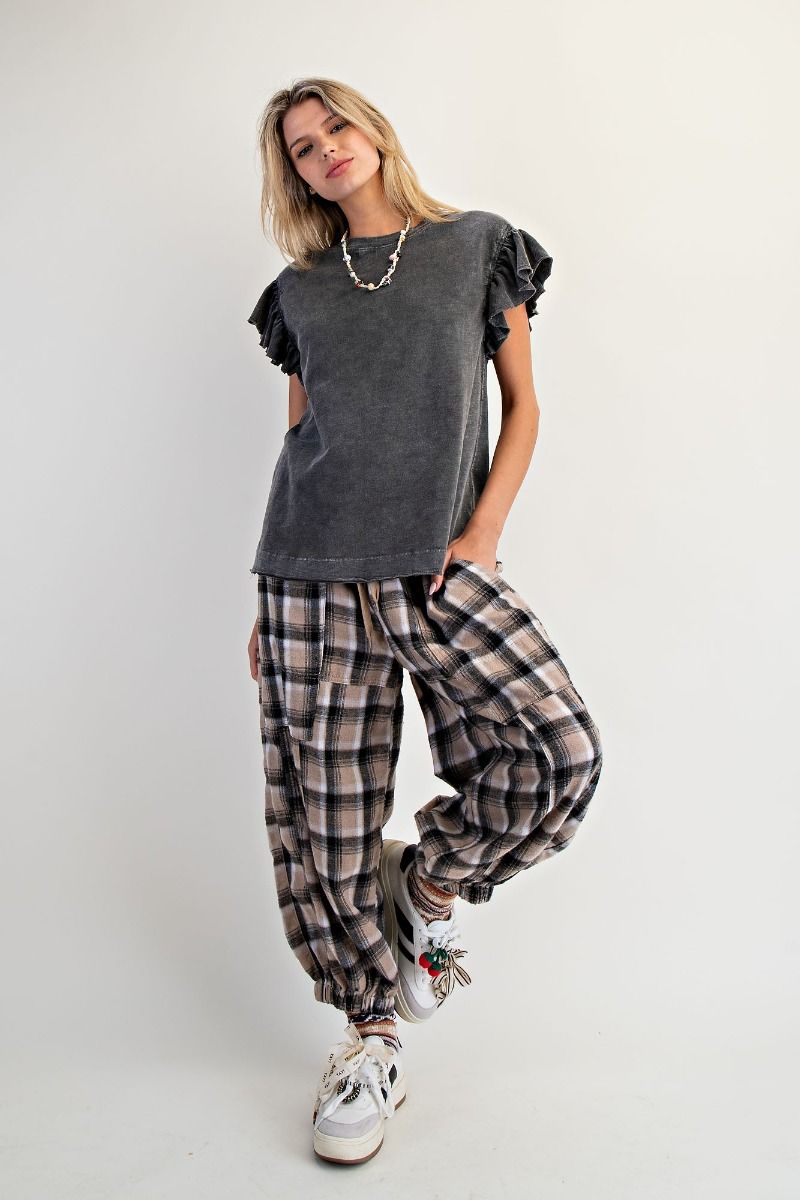 A woman modeling a dark gray short-sleeve t-shirt with ruffled sleeves, paired with beige and black plaid harem pants and white sneakers, against a plain light gray studio background.
