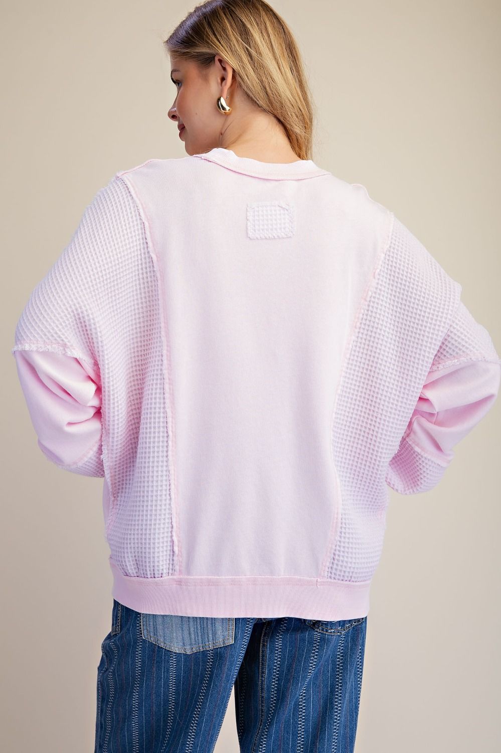 A woman shown from the back wearing a light pink textured sweatshirt with waffle-knit sleeves and a small rectangular patch on the upper back, paired with blue jeans against a plain beige studio background.