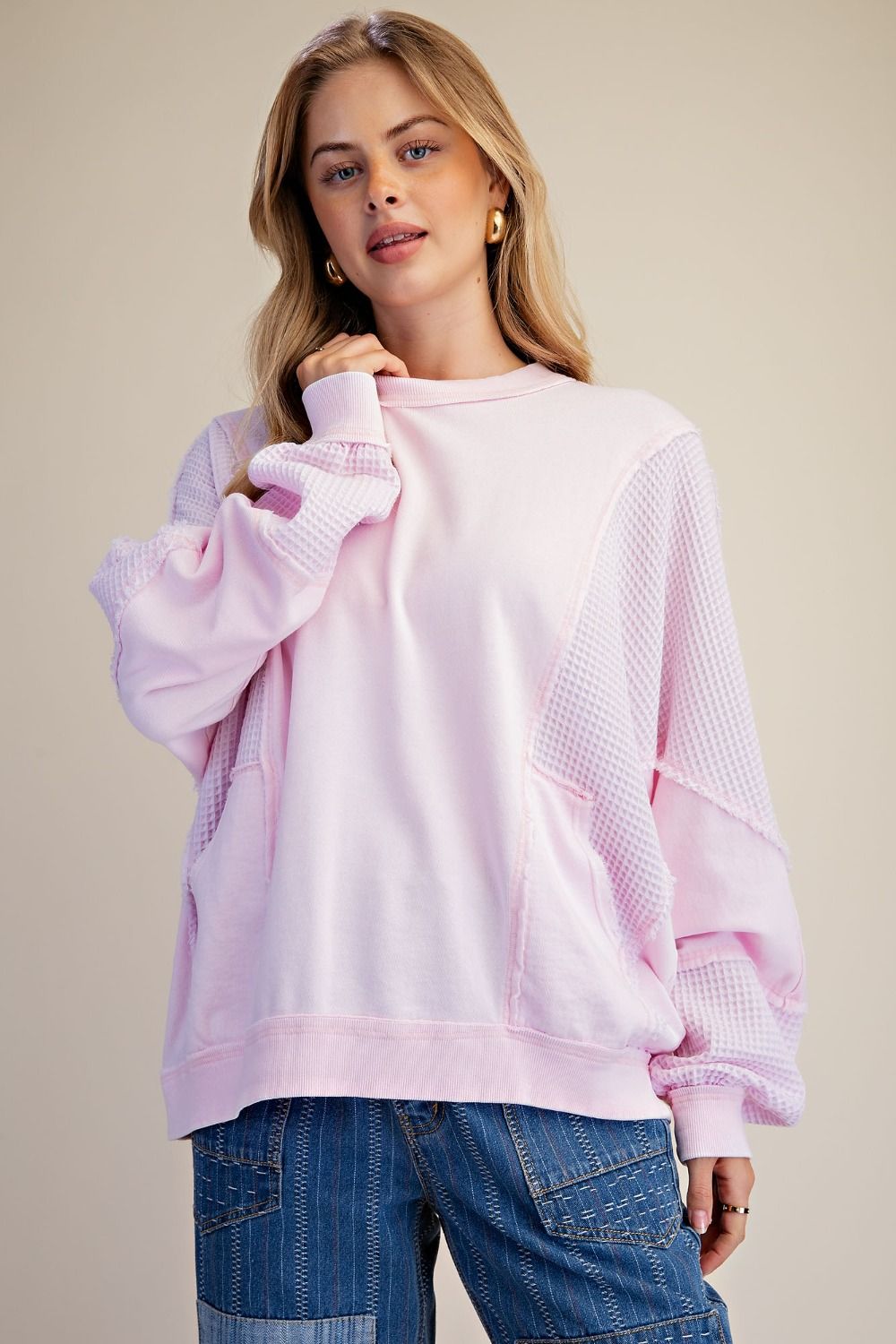 A woman wearing a light pink crewneck sweatshirt with textured waffle-knit side panels and blue jeans, posed against a solid beige background.