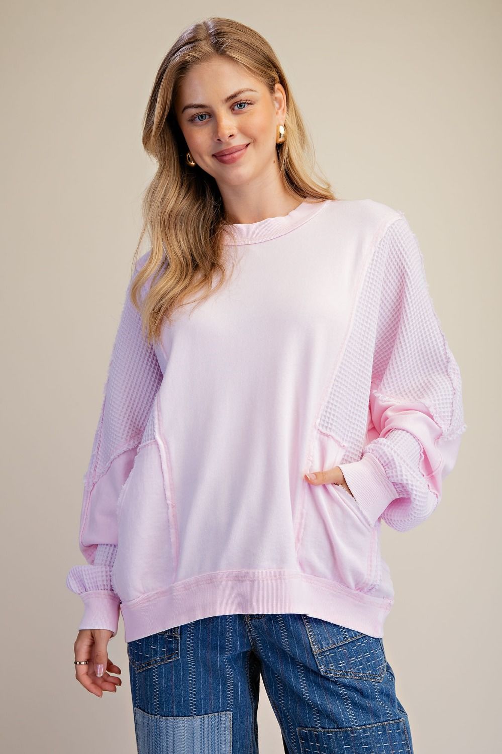 A woman wearing a light pink crewneck sweatshirt with textured waffle-knit sleeves and side pockets, paired with blue jeans, against a solid beige background.