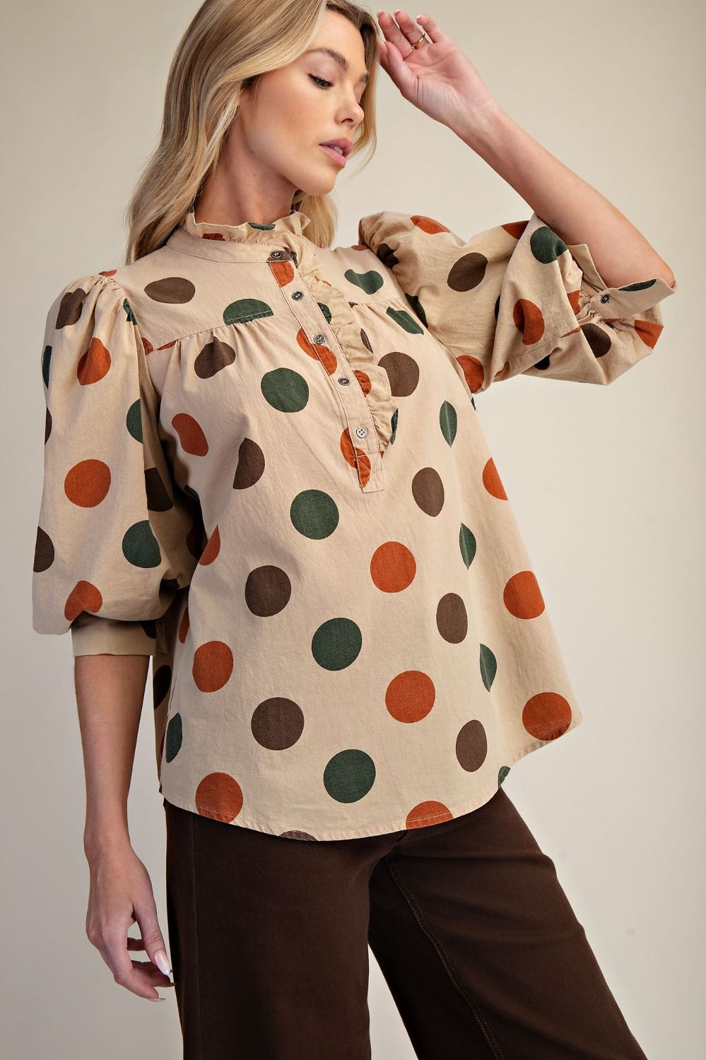Woman wearing a beige blouse with colorful polka dots and brown pants against a neutral background