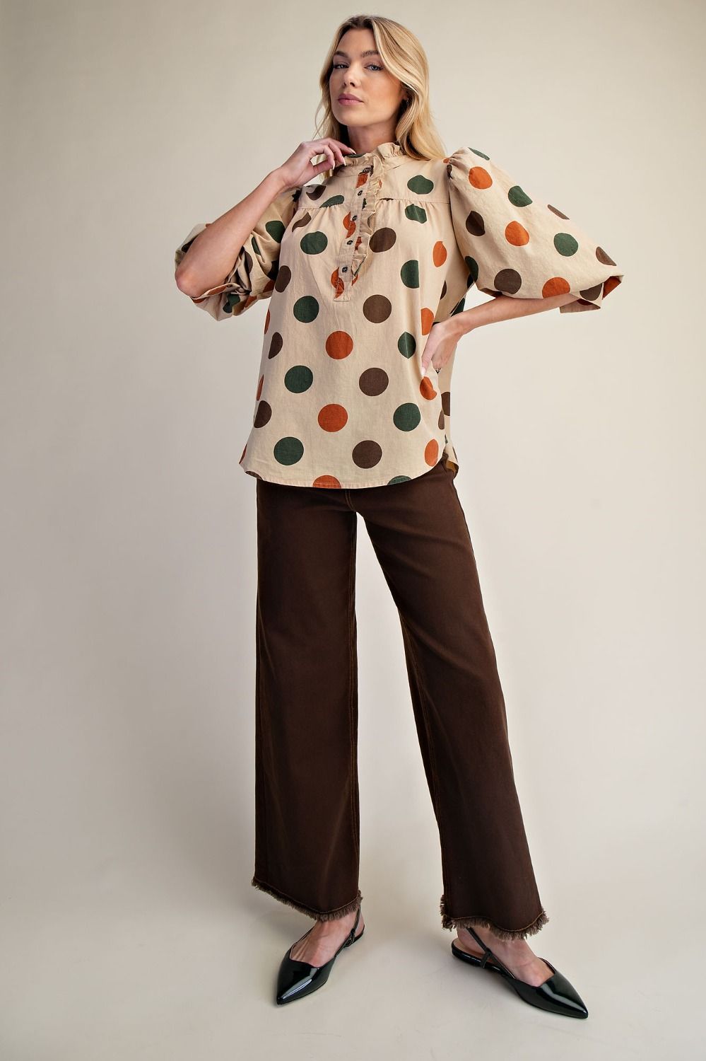 A woman modeling a beige blouse with large multicolor polka dots in brown, orange, and green, paired with dark brown wide-leg trousers and black pointed flats, against a plain beige studio background.
