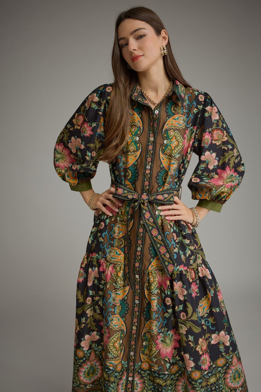 A woman modeling a long-sleeved button-front dress with a tie waist and an intricate paisley and floral print in dark green, gold, pink, and black, set against a plain gray studio background.