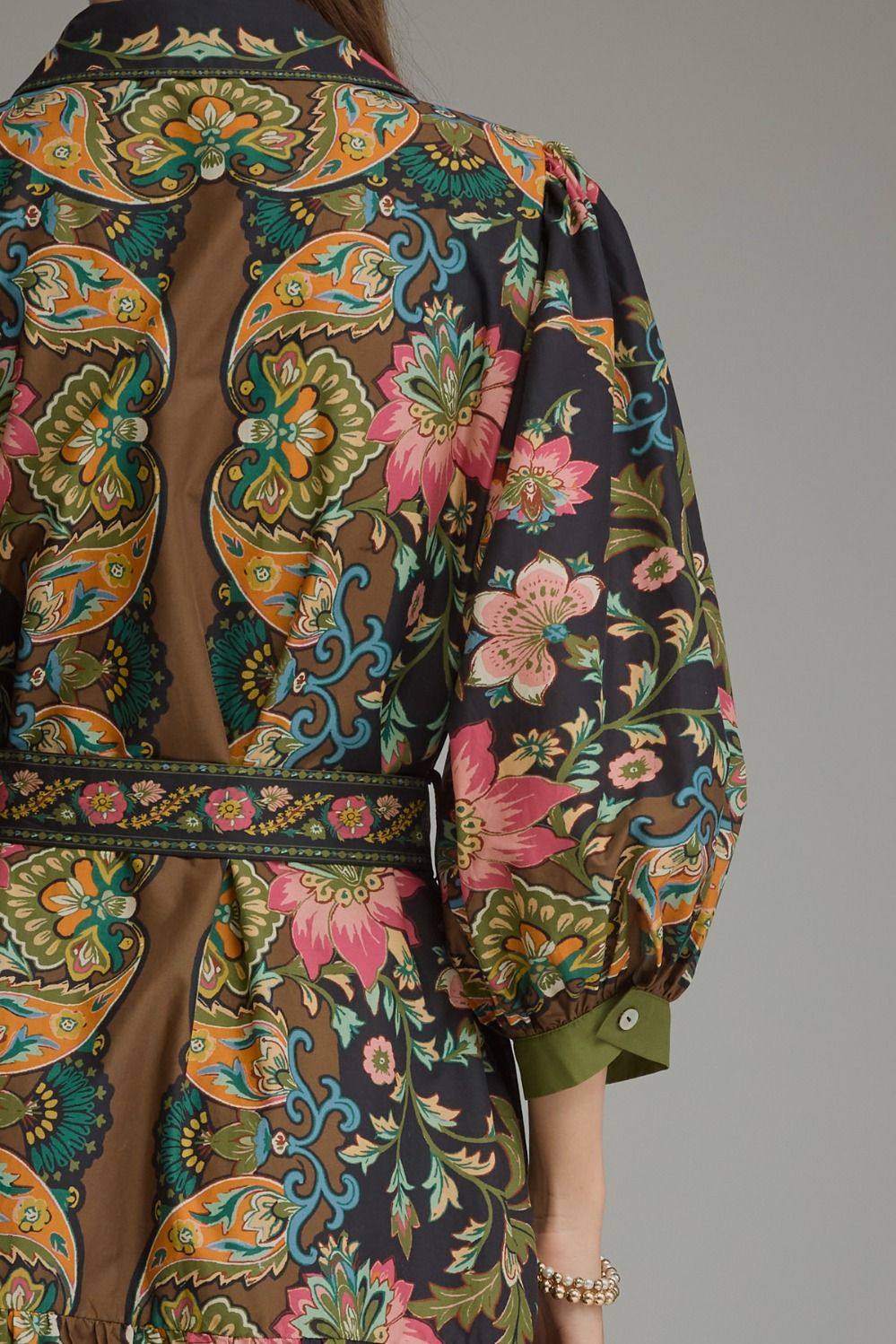 Close-up back view of a person wearing a long-sleeve patterned robe or kimono-style garment with a brown base and ornate paisley and floral print in orange, teal, pink, and green. The back has a large open panel and a matching patterned waist tie.