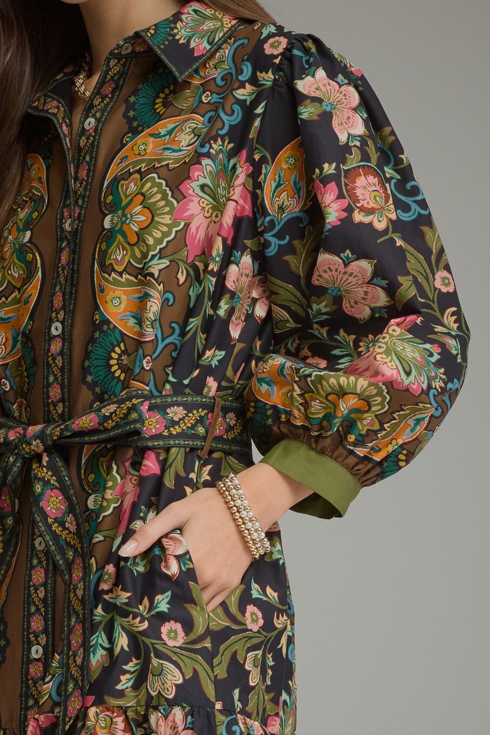 A close-up of a woman wearing a long-sleeve collared shirt dress with a dark base and a vibrant multicolor paisley and floral print. The dress features an olive green cuff, a matching waist tie, and a sheer mesh lining.