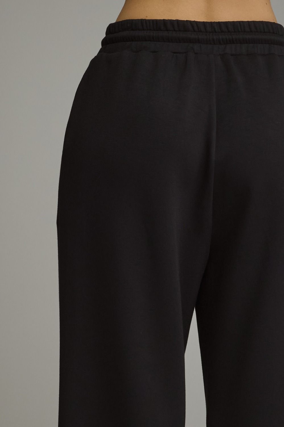 Close-up of a person wearing black wide-leg sweatpants, showing the elastic waistband and side seam against a plain light gray background.