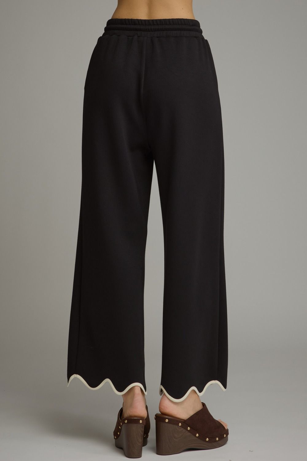 Black wide-leg cropped pants with a scalloped white hem, shown from the back on a model wearing brown wedge sandals against a neutral gray background.