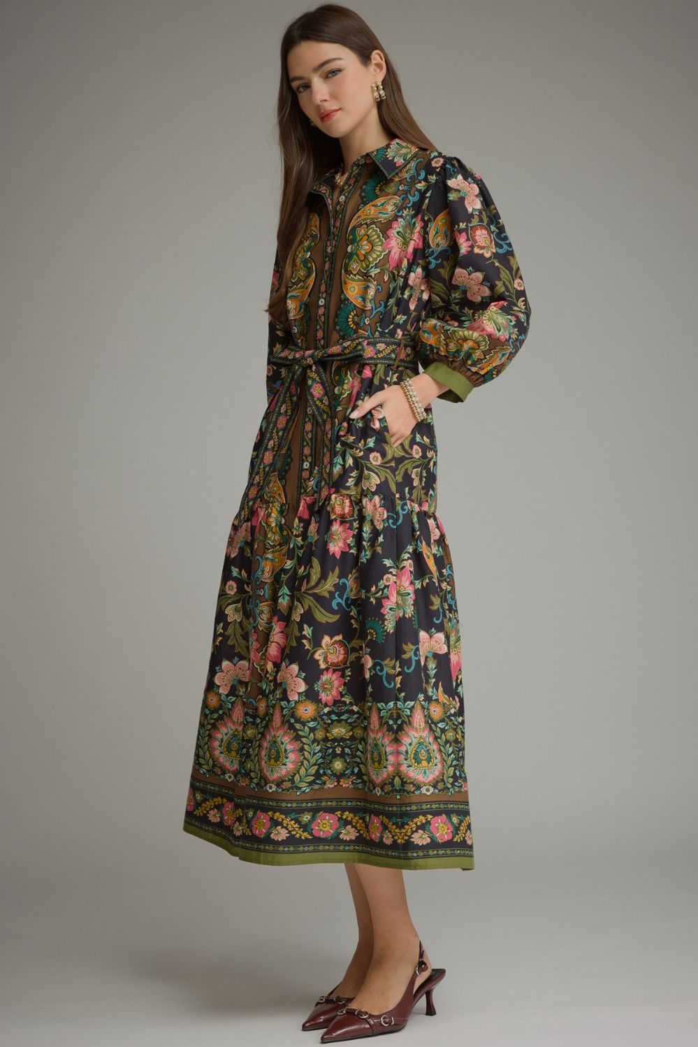 A woman models a long-sleeved midi dress with a dark navy base and an intricate multicolor floral and paisley print. The dress features a tie-neck detail, a matching fabric belt, and a decorative border along the hem.
