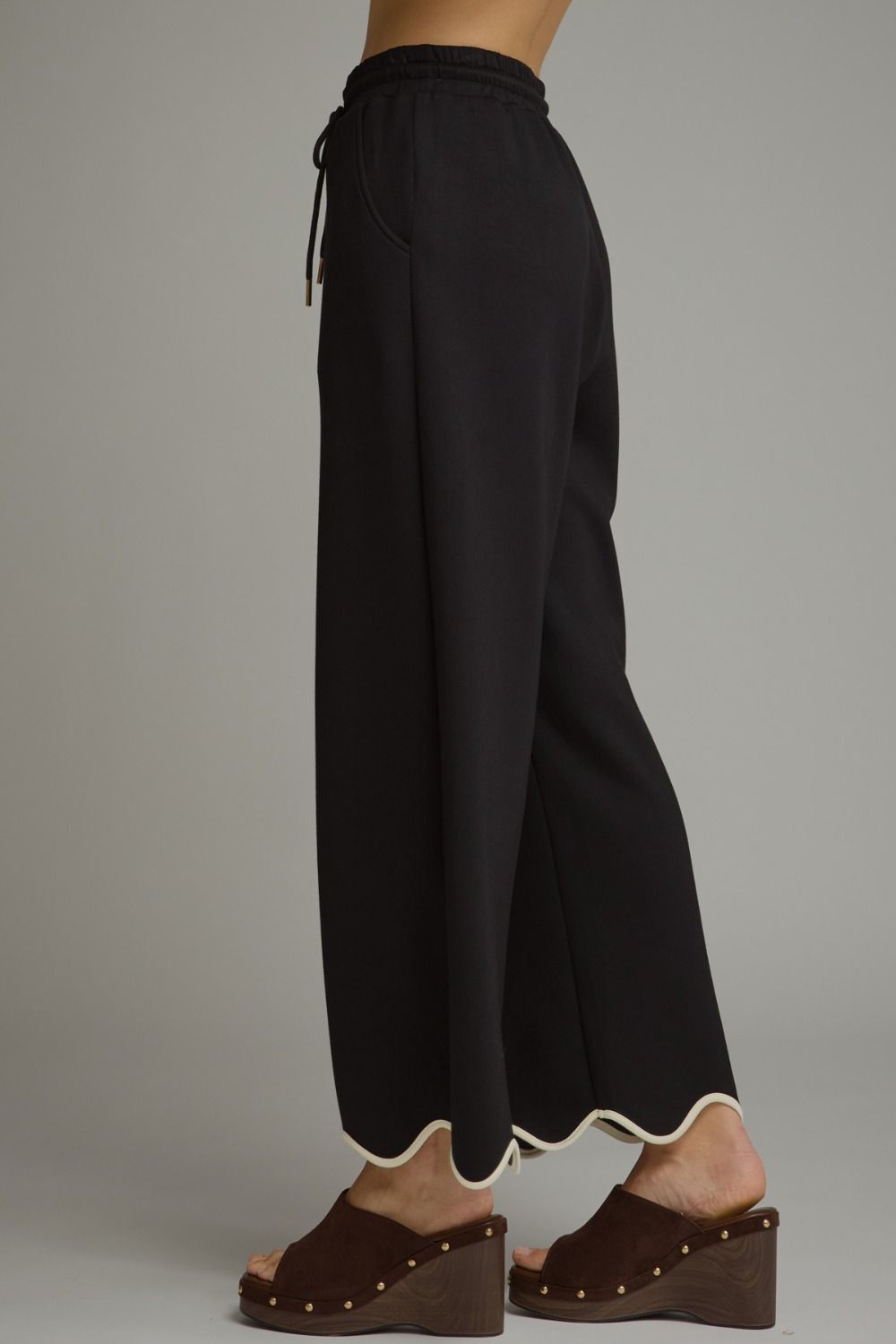 Black wide-leg cropped pants with a scalloped hem and drawstring waist, shown on a model wearing brown wedge sandals against a neutral gray background.
