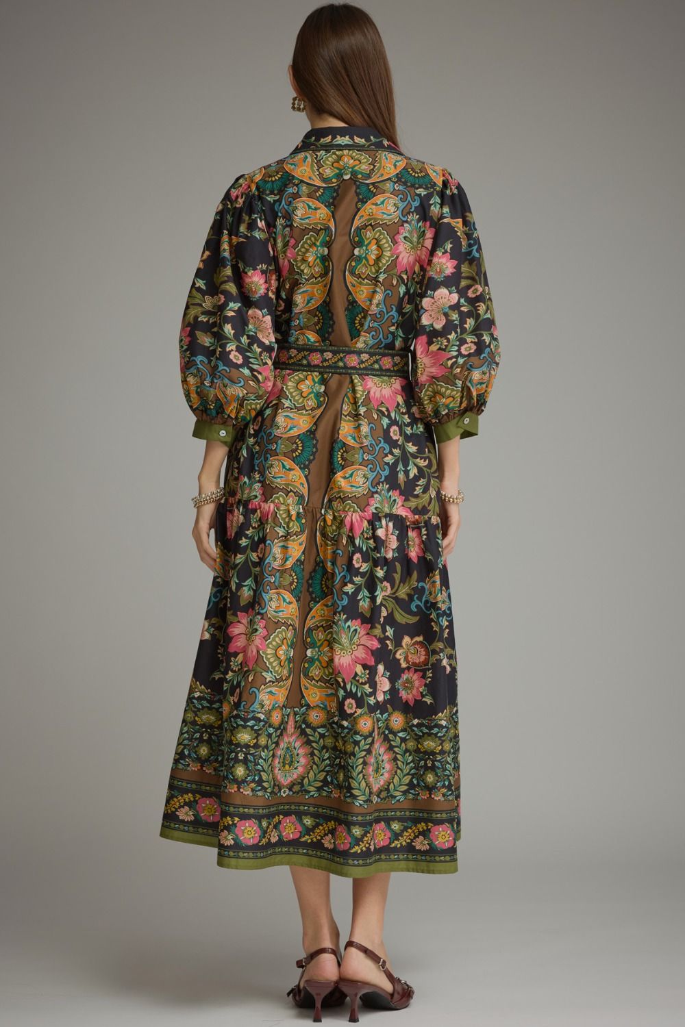 A woman viewed from the back wearing a long-sleeved midi dress with a dark base and an intricate multicolor floral and ornamental print. The dress features a matching belt and is paired with dark brown strappy heels against a solid gray studio background.