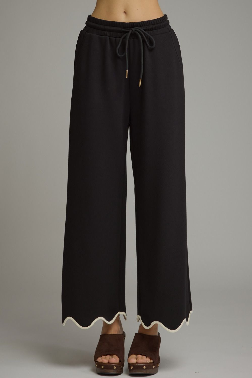 Black wide-leg cropped pants with a drawstring waist and scalloped hem, shown on a model wearing brown platform sandals against a gray studio background.