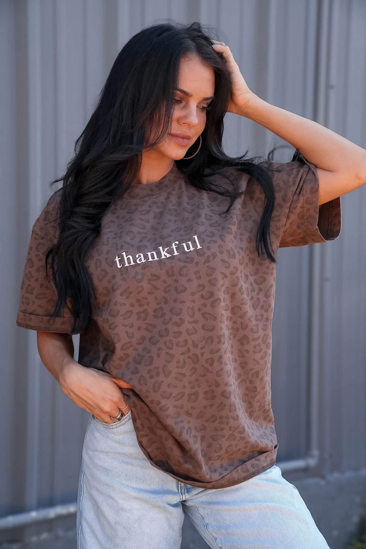 A woman wearing a brown leopard-print t-shirt with the word 'thankful' printed in white across the chest, paired with light-wash jeans, posing in front of a gray corrugated metal wall.