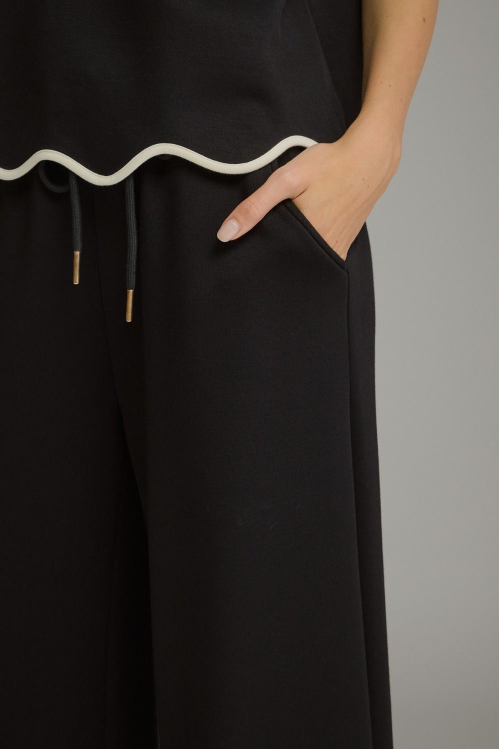 Close-up of a person wearing black wide-leg trousers with a white wavy trim detail at the waistband and black drawstrings with gold-toned tips, with one hand in the pocket.