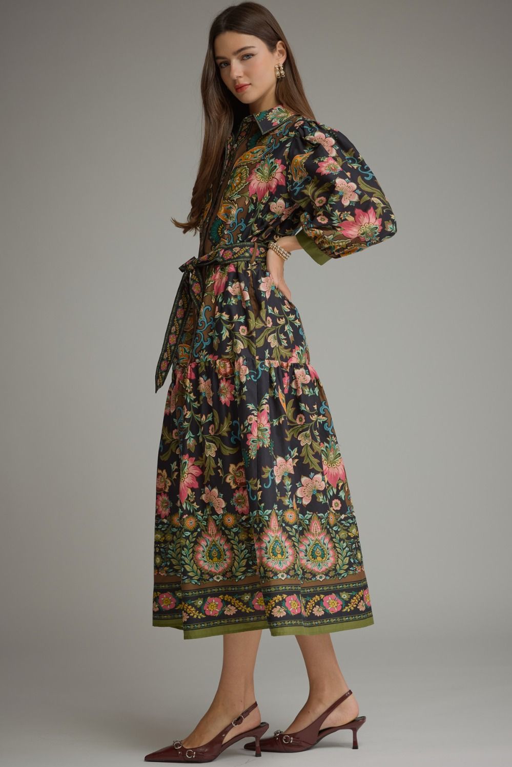 A woman models a high-neck midi dress with a dark background and a vibrant, multicolored floral and paisley print. The dress features voluminous sleeves, a matching waist tie, and a decorative border at the hem, paired with dark brown slingback heels against a neutral gray studio background.