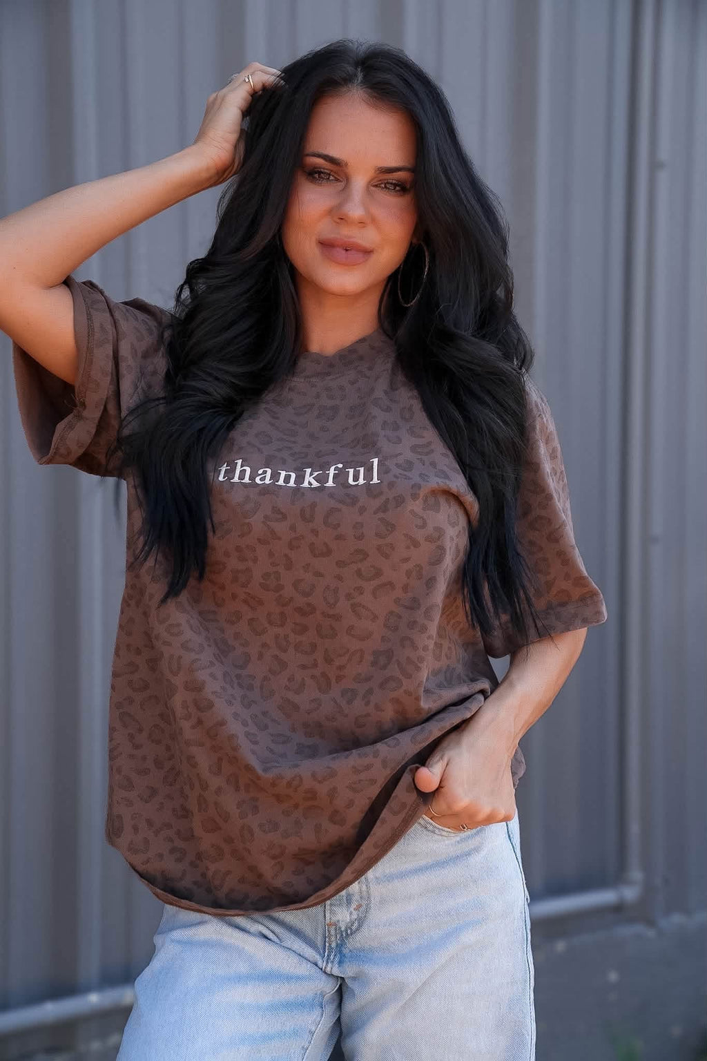 A woman wearing a brown leopard-print t-shirt with the word 'thankful' printed in white across the chest, paired with light-wash jeans, posing against a gray corrugated metal wall.