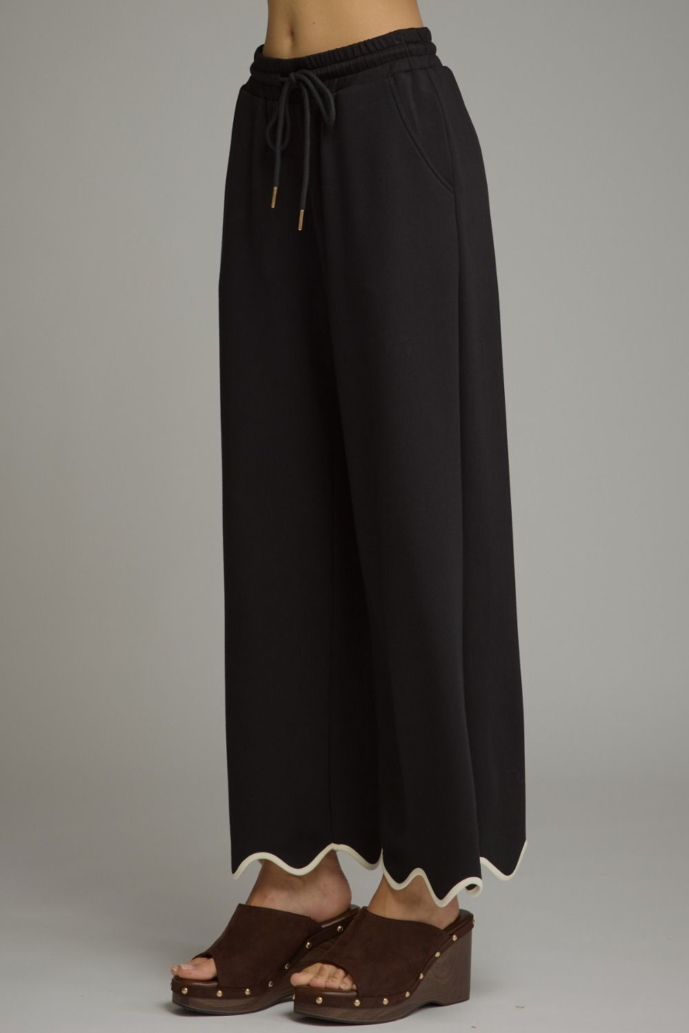 Black wide-leg cropped pants with a drawstring waist and scalloped hem, shown on a model wearing brown wedge sandals against a neutral gray background.