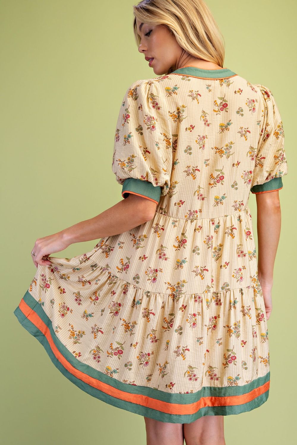A woman wearing a cream-colored tiered dress with a small floral print, featuring sage green and orange striped trim, shown from the back against a solid light green background.