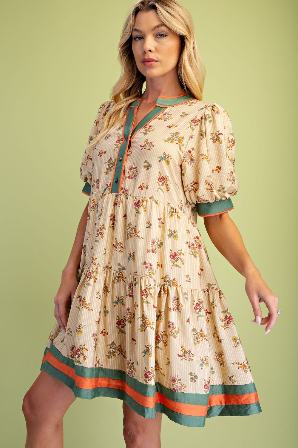 A woman modeling a short-sleeve tiered dress with a beige floral print and contrasting teal and orange trim, set against a solid light green background.