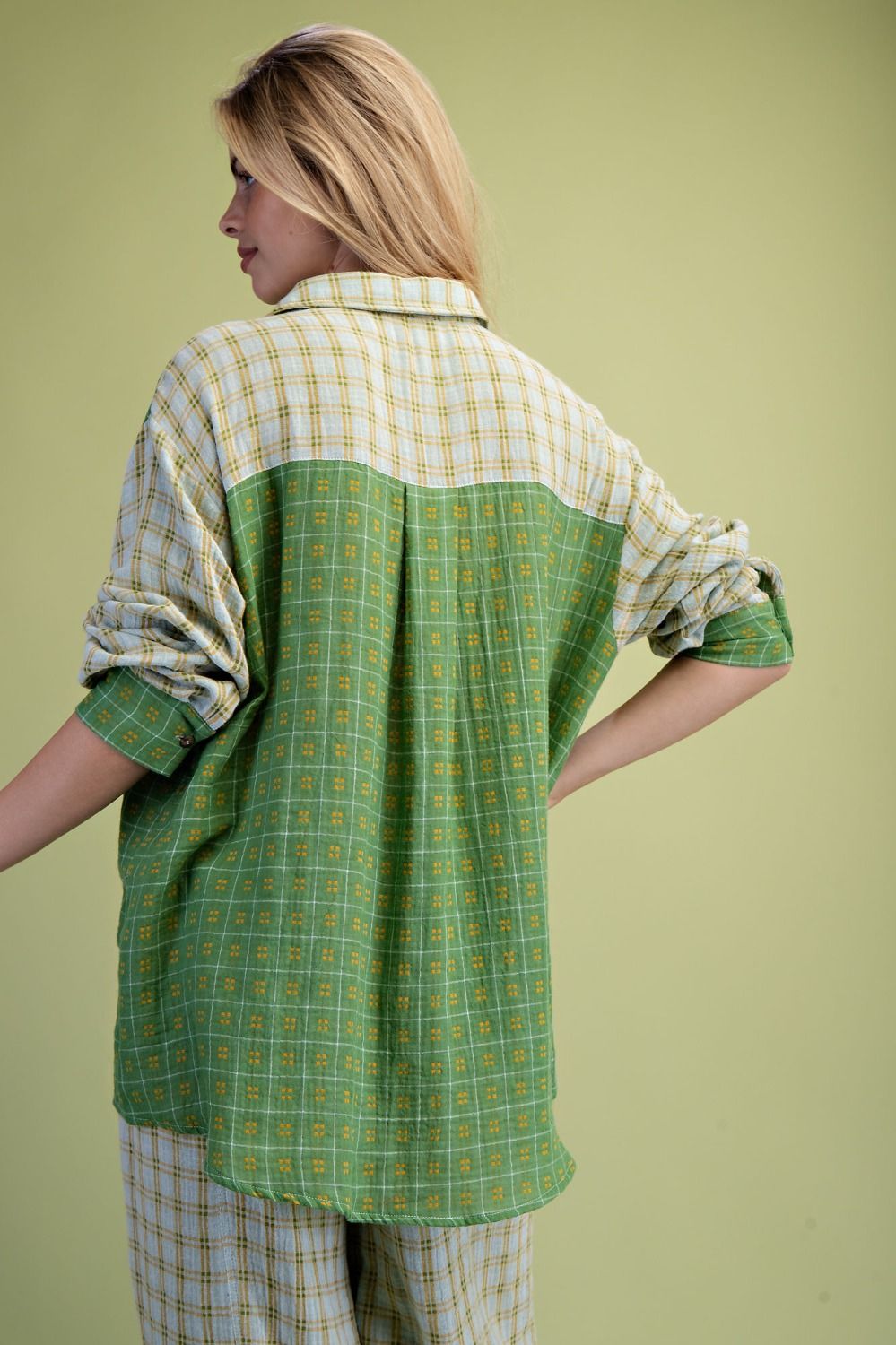 A woman shown from the back wearing a loose button-up shirt with a color-blocked plaid design, featuring a green and yellow lower half and a cream and yellow upper half, paired with matching plaid pants against a solid light green background.