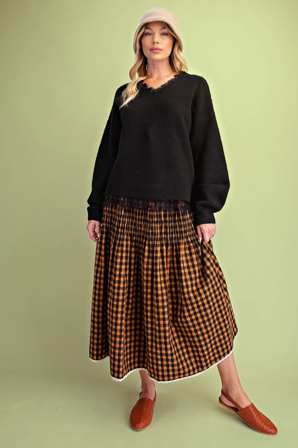 A woman modeling a black knit sweater and a brown and black checkered midi skirt with white trim, paired with brown leather mules and a pink beanie, against a solid light green background.