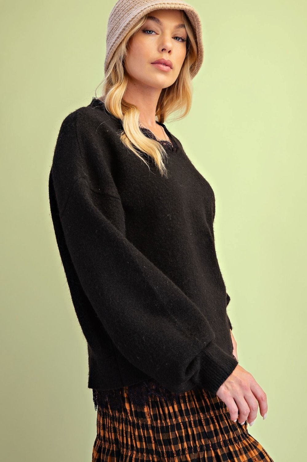 A woman wearing a dark brown fuzzy knit crewneck sweater, a beige knit beanie, and an orange and black plaid skirt, posed against a solid pale green background.