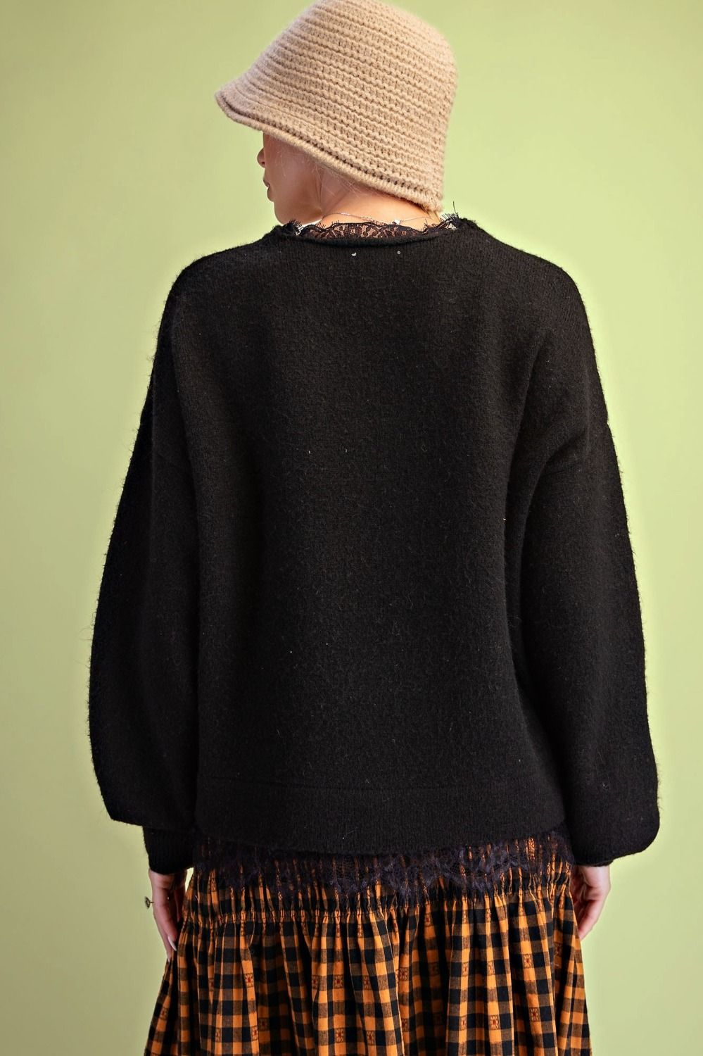 A person shown from the back wearing a loose black knit sweater, a beige bucket hat, and an orange-and-black plaid skirt against a pale yellow-green studio background.