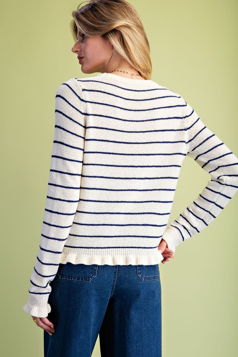 A woman shown from the back wearing a cream knit sweater with thin navy horizontal stripes and blue jeans, standing against a solid pale yellow-green background.