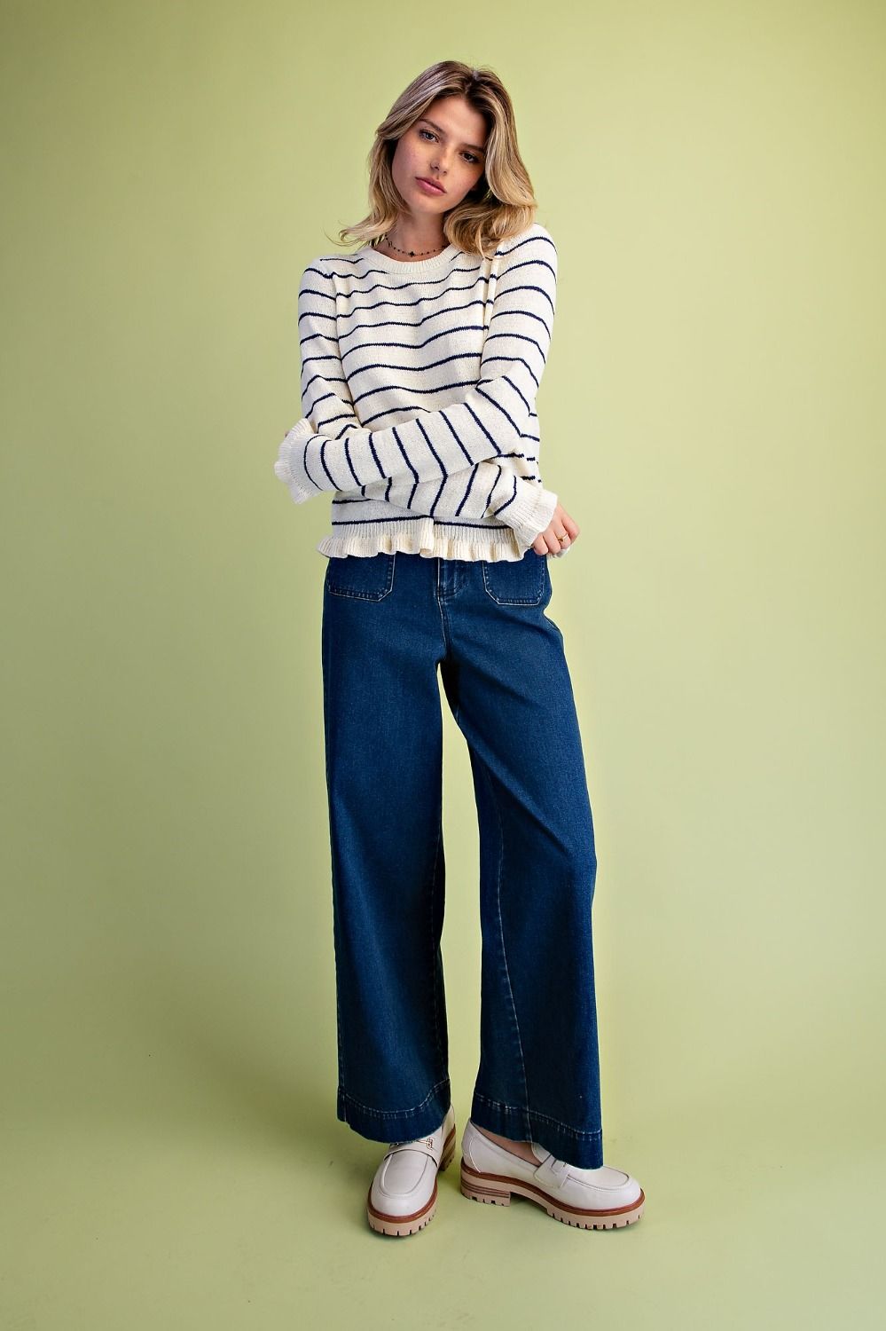 A woman modeling a white long-sleeve knit sweater with thin navy horizontal stripes, paired with dark blue wide-leg jeans and white platform shoes, standing against a solid pale green studio background.