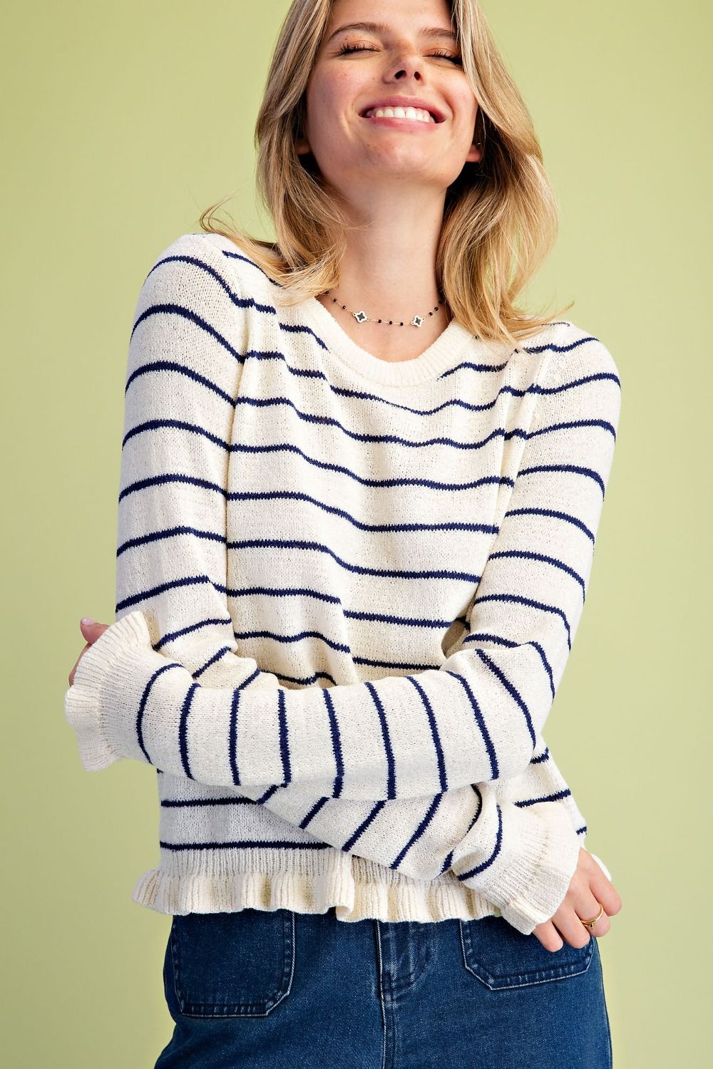 A woman wearing a cream knit sweater with navy blue horizontal stripes, paired with blue jeans, set against a solid light olive green background.