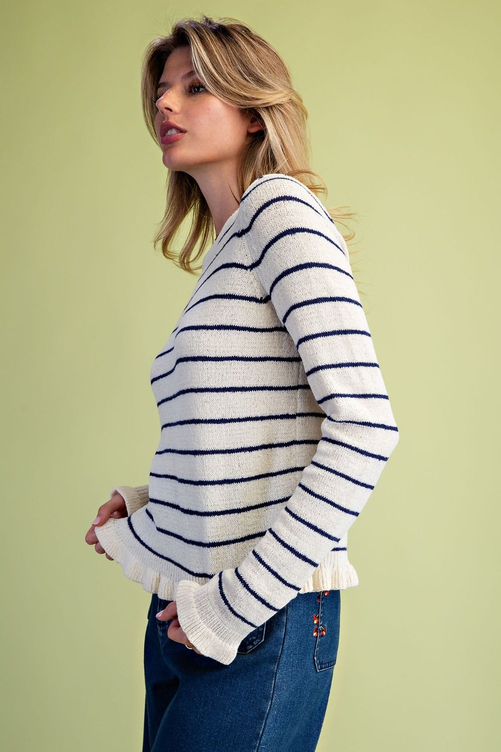 A woman wearing a cream knit sweater with navy blue horizontal stripes and blue jeans, standing against a solid pale yellow-green background.