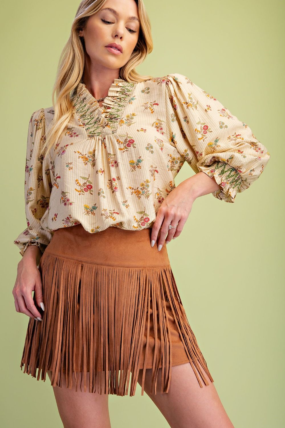 A woman modeling a cream floral long-sleeve blouse with ruffled trim and a brown fringe mini skirt against a solid light green background.