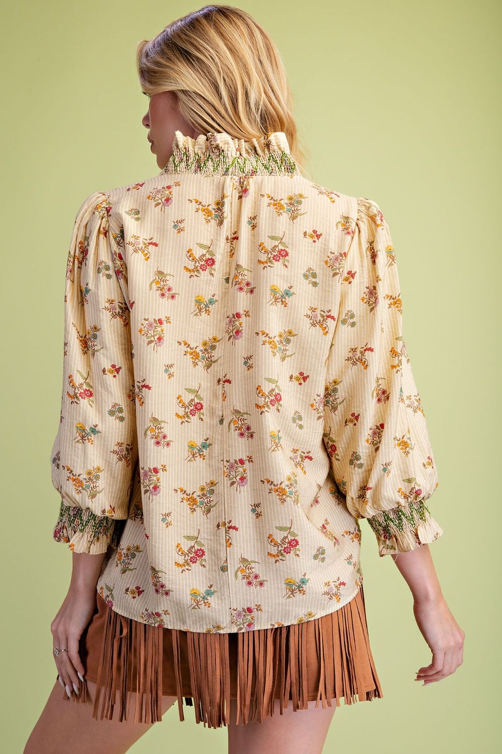 A woman shown from the back wearing a cream-colored, textured blouse with a small multicolor floral print, puffed sleeves, and a ruffled high collar, paired with a brown fringe skirt, against a solid light green background.