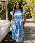 Woman in a blue floral dress standing outdoors on a wooden path with greenery.